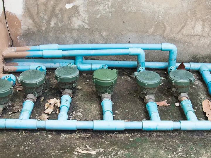 Fileira de hidrômetros verdes instalados em tubulação azul, no chão de um condomínio
