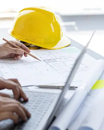Pessoa desenhando um projeto com régua e notebook, ao lado de um capacete amarelo